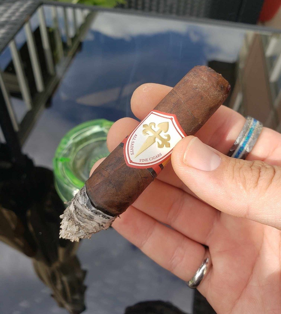 Saint Francis All Saints cigar is chocolatey brown with a white and gold shield emblem on the band. In this picture it has been smoked until the final third, right under the band