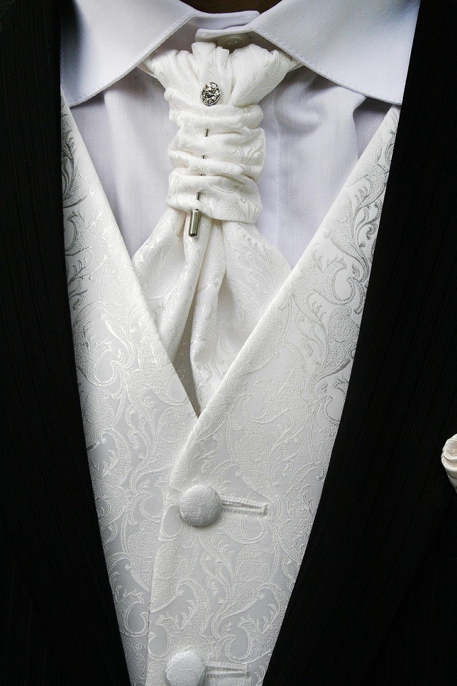 A man in a black tuxedo is wearing a solid white formal ascot with a silver tie pin holding the knot together. He is also wearing a plain white dress shirt and a white vest with a light paisley pattern