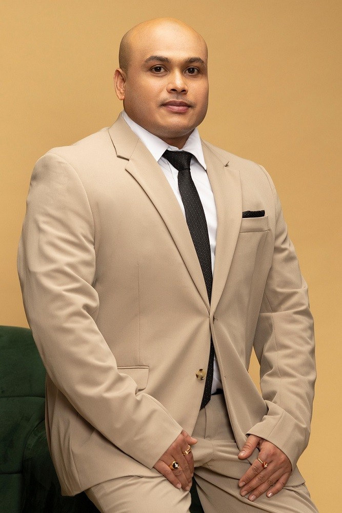 A large man is wearing a light brown suit and white dress shirt. He also has a skinny black tie and matching black pocket square
