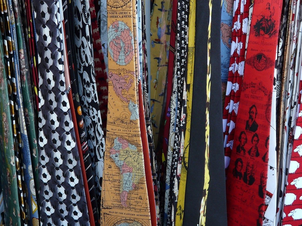 Numerous novelty ties are hanging off a tie rack. The patterns range from scoccer balls to fish to maps of the world to butterflies, and more. They are all very colourful and bold