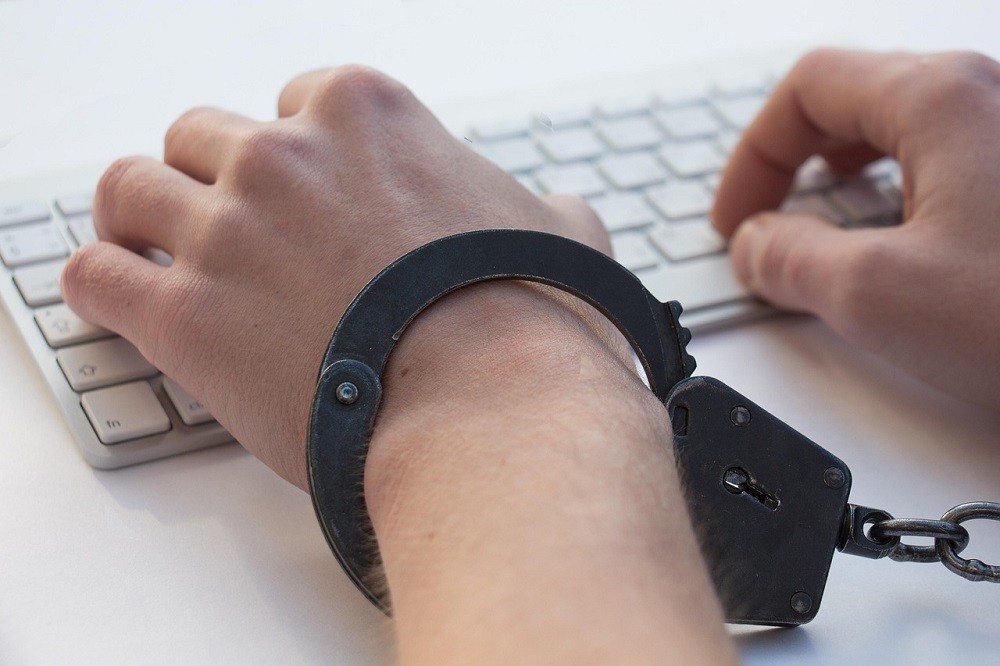 A man is typing on a keyboard. He has handcuffs on, locking him to the keyboard