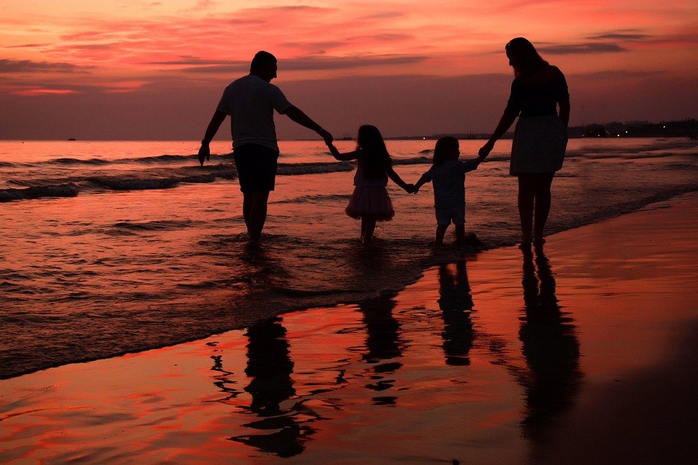 A father, mother, and two children are walking along the beach at sundown. The picture has vibrant hues of purples, reds, and oranges