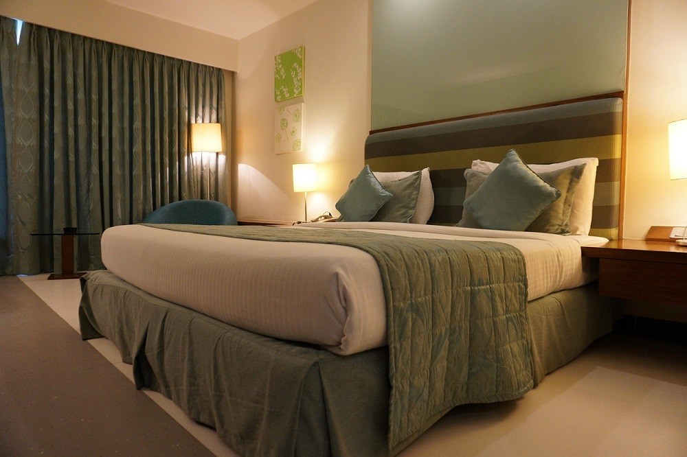 A hotel room with a large, king-sized bed with several pillows and blankets. All in light shades of greens and blues