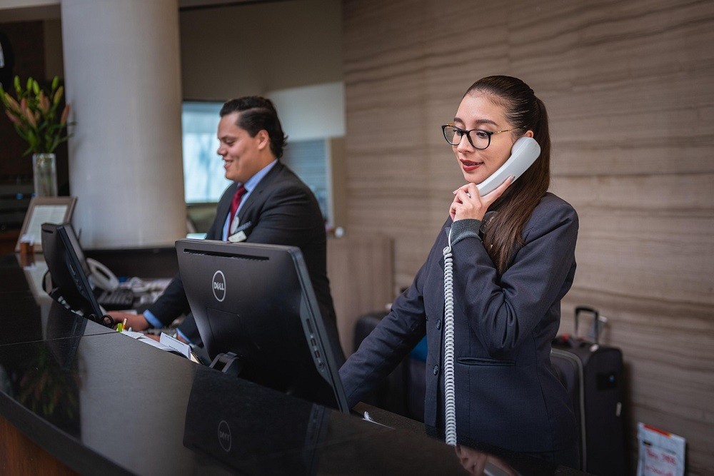Two hotel receptionists in black suits (one male, one female) are working at the front desk at a hotel. The female is on the phone while the male looks at the desktop