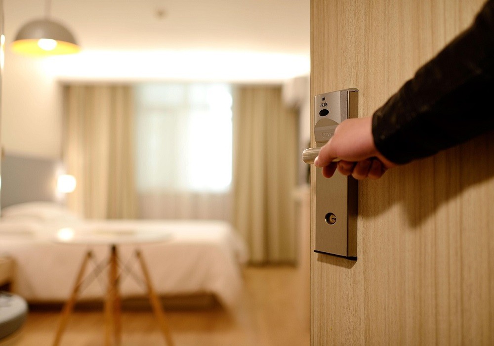 A man is opening the door to his hotel room. A white bed and small, wooden table can be seen