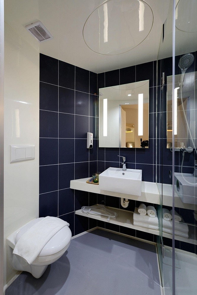 A hotel bathroom has a dark blue tile backsplash, white toilet and sink, and white towels rolled up on a shelf under the sink