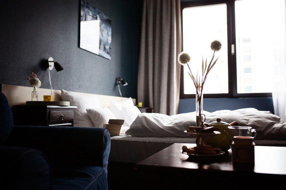 A hotel room shows a small vase with two purple flowers sitting on a dark brown table. The walls of the room are dark blue and the bed has white sheets