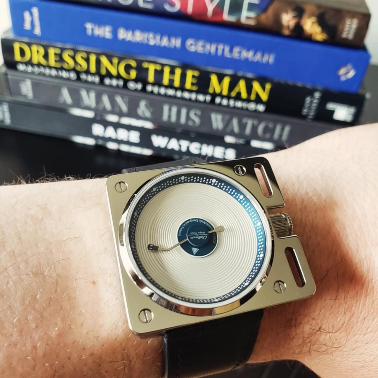 A wrist watch shaped like a vinyl record player has a white vinyl record with blued hands on it inside a shiny silver square case and a black strap