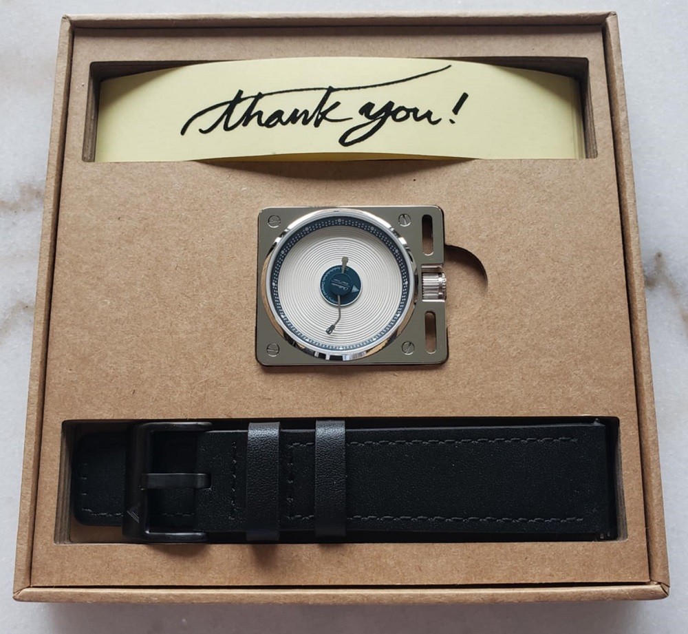 The inside of the Esteemed Gentleman vinyl record box contains a handful of black and white stickers with AndoAndoAndo printed on it in a variety of fonts, the watch itself nested in the middle of the box, and a slot for the black leather strap near the bottom of the box. The box itself is light brown in colour and square in shape