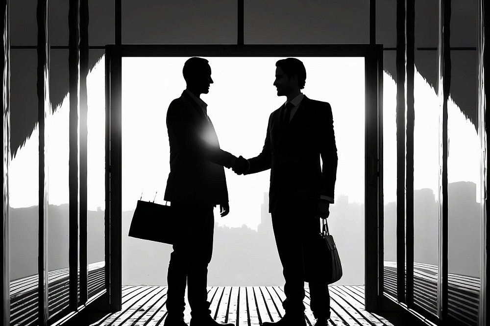 Two businessmen are standing in a hallway shaking hands. Both of them are younger, clean cut, and dressed in black suits