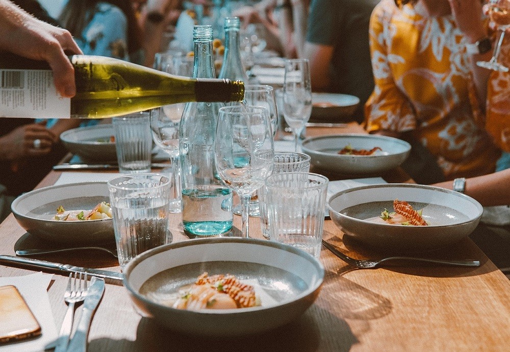 A large place-setting has various types of silverware, glasses, and plates on it. The food is just being dished out and wine is being poured into a glass