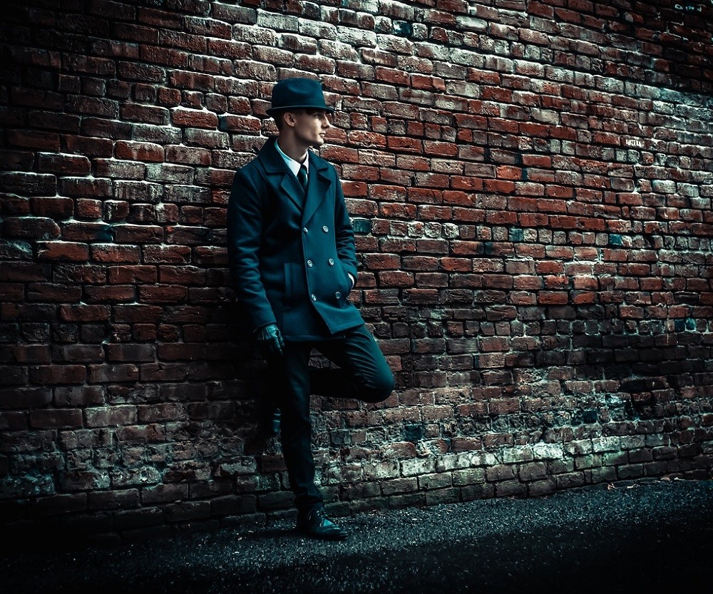 A middle-aged man in a tailored navy double-breasted suit, matching fedora, and dark brown dress shoes leans against a brick wall