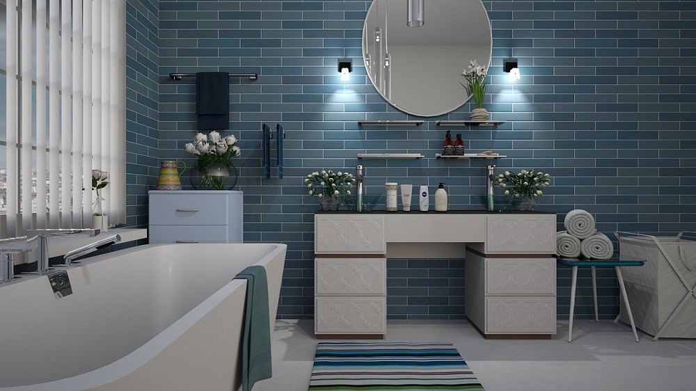 A bathroom with blue tile and a large round mirror has a white bathtub, white counters, and a sink with a soap dispenser beside it