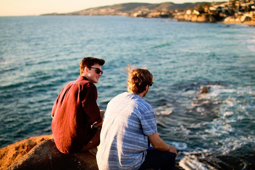 Two young men sit on a rock on the shore of a lake. They are having a conversation
