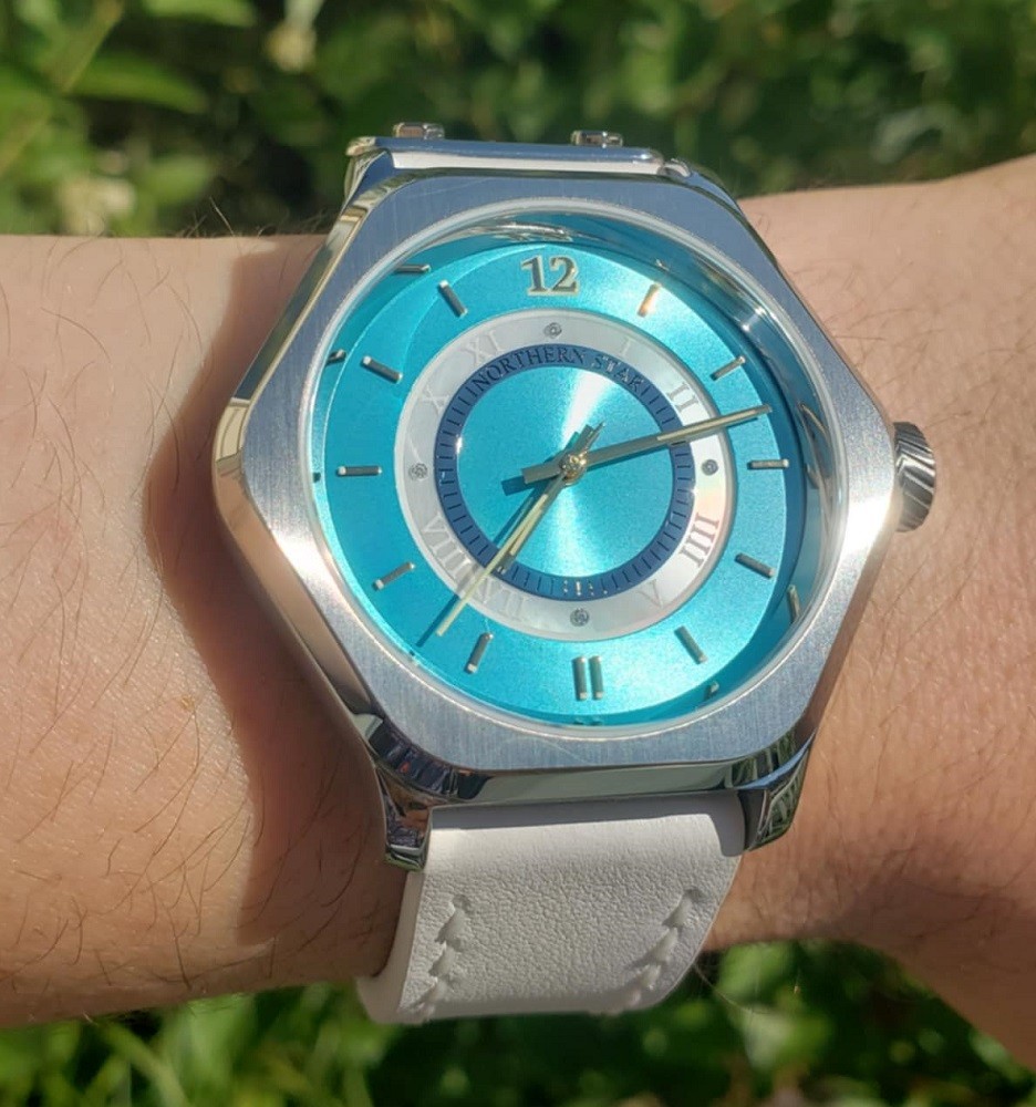 A six-sided silver watch features a bright teal dial with gold markers. The watch is on a white leather strap
