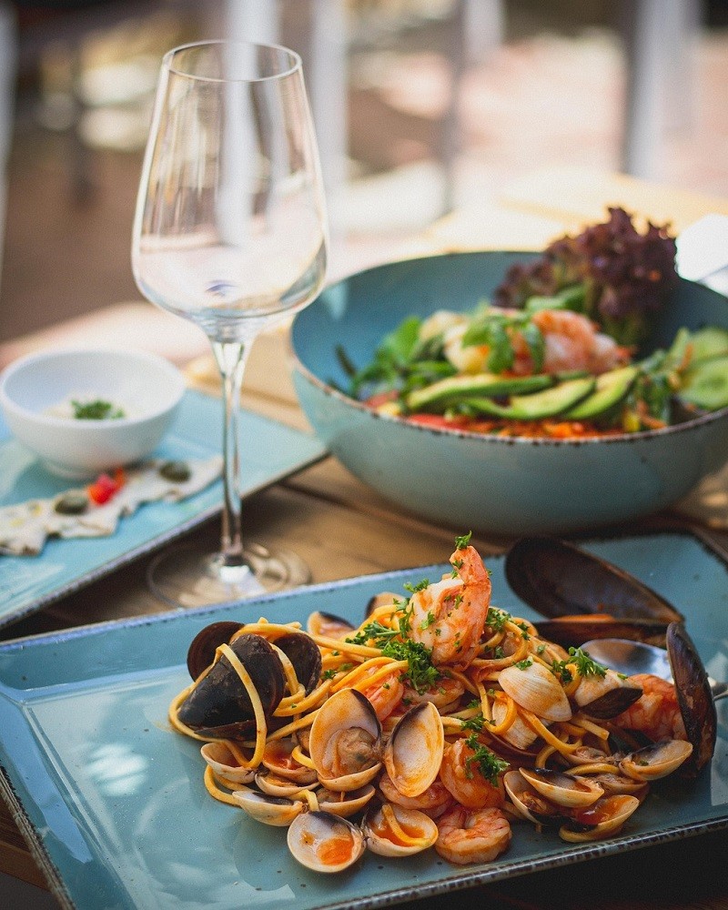 A seafood meal is plated on a blue square plate. The meal has pasta like spaghetti with clam shells and red tomato sauce on it. There is a salad beside it with various vegetables and leafy greens inside
