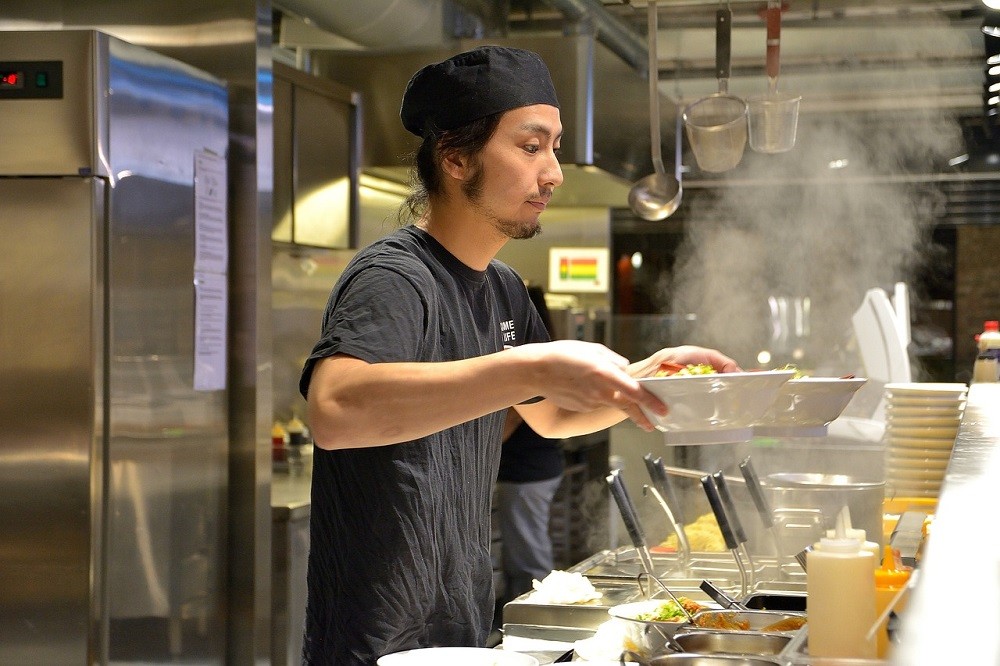 A chef wearing all black holds two bowls in his hands. He is placing them where a server can easily grab them