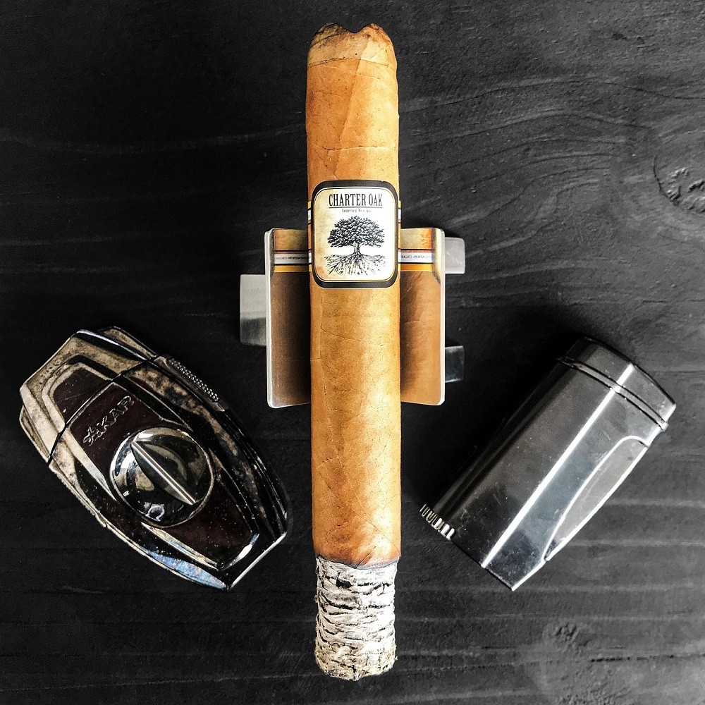 A light brown cigar sits on a silver cigar holder. There is a silver V cutter to the left of the cigar and a silver lighter to the right