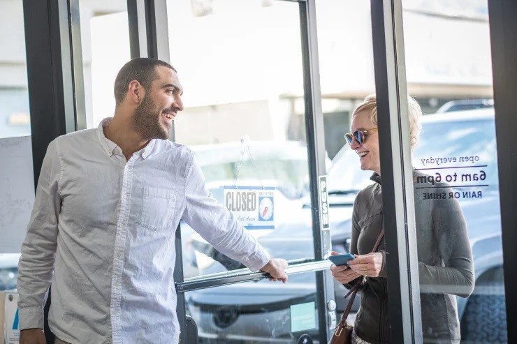 A man in a white dress shirt holds a door open for a woman wearing a grey sweater who is walking into the building