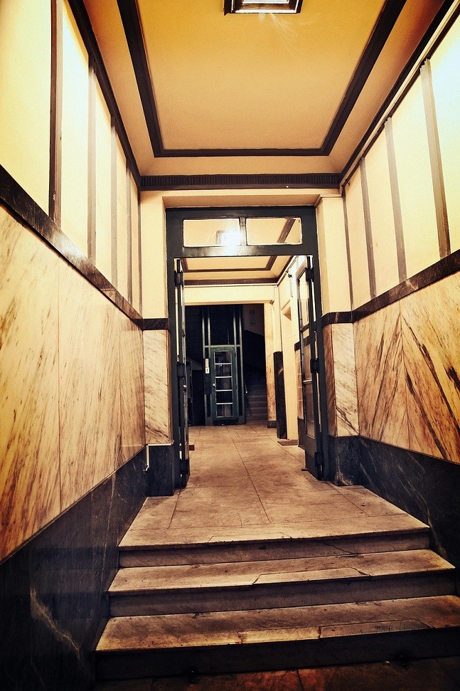The interior of a hallway of a common area at an apartment building. The walls are yellow with black trim and the floor is marble. There is a single black door at the end of the hallway