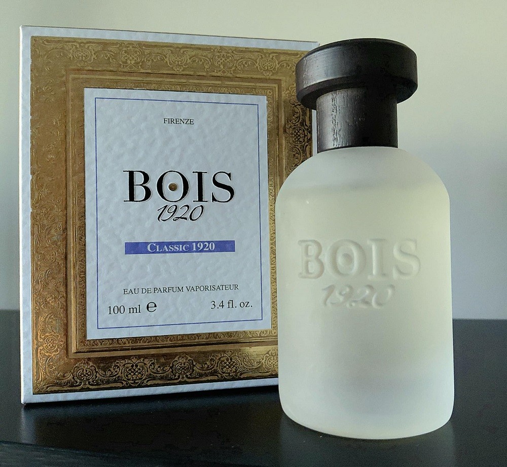 A clear bottle of Bois 1920 fragrance has a black cap. It sits beside its presentation box that has a large gold border with a white inner part with the name of the fragrance and manufacturer inside