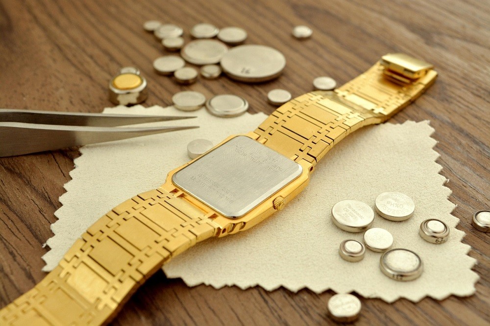 A gold quartz watch sits on a table surrounded by various shapes and sizes of watch batteries