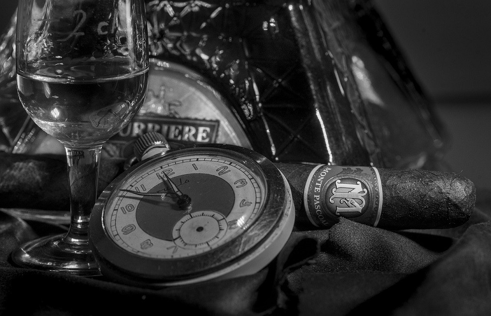 A black and white image of a pocket watch, lit cigar, and small glass of liquor