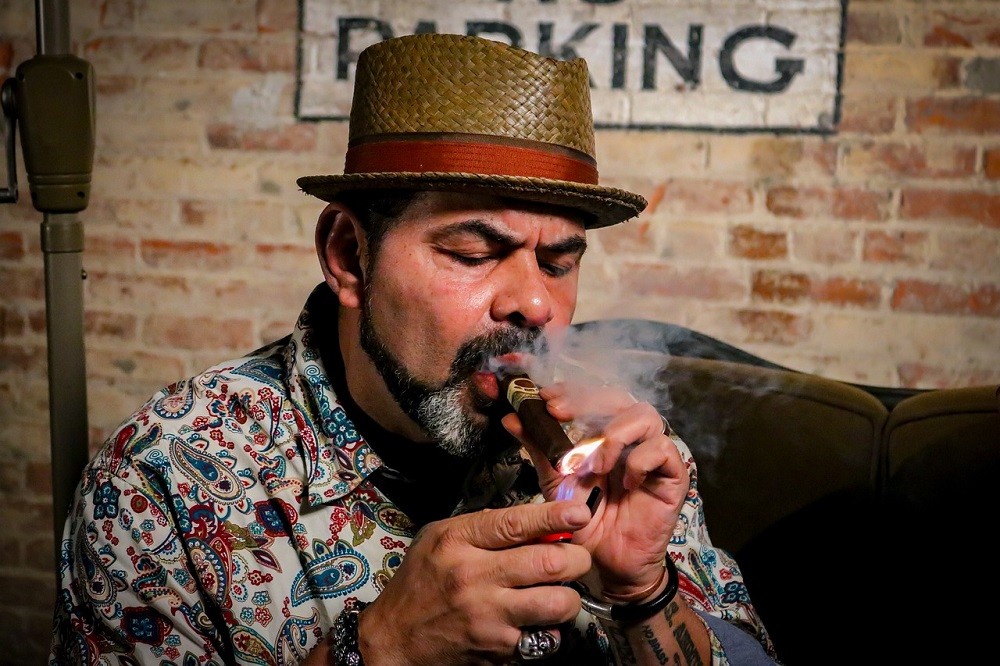 A man wearing a straw hat is lighting a cigar with a soft flame lighter. Smoke is coming out of the lit end of the cigar