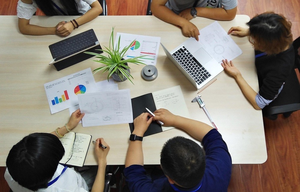 A group of five young white-collar employees sit at a desk with various spread sheets, notes, and charts on the table. They are pointing and discussing one of the charts