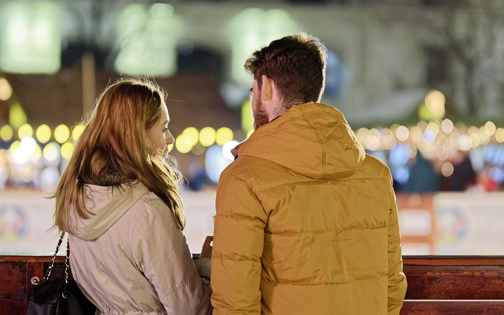 A young man and woman are sitting on a bench. They are facing each other and in mid-conversation