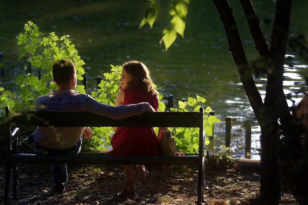 A young couple are sitting on a bench facing a river. The man has his arm around the woman as they both look at the river