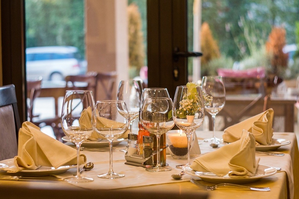 A table for four people at a fancy restaurant is set. There are white plates with white cloth napkins laid out on them, wine glasses, and cutlery on each side of each plate
