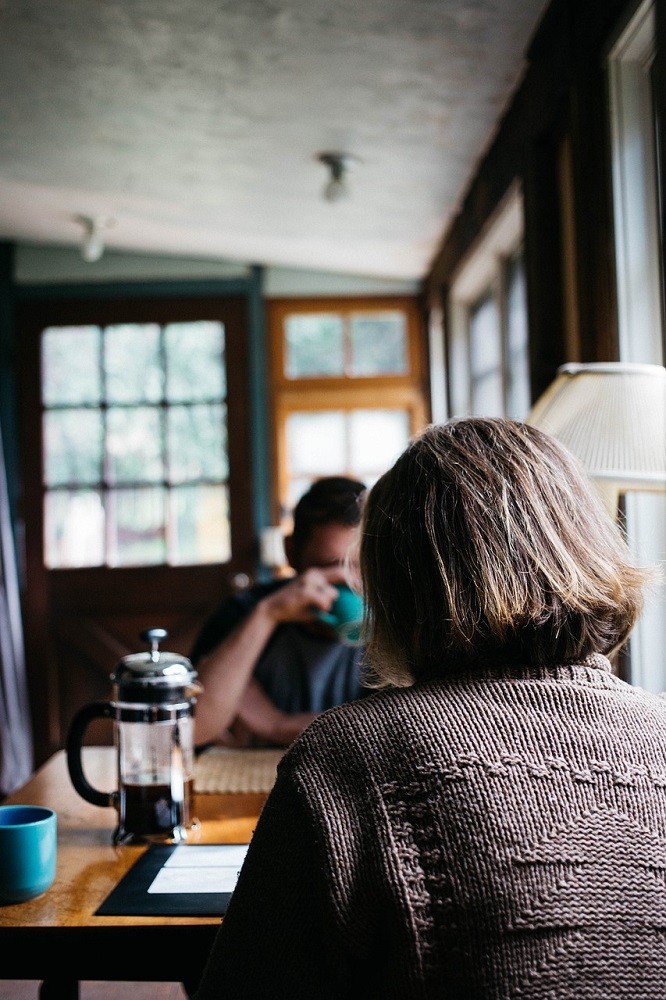 A man and woman sit at a table in a coffee shop. The man is sipping from his mug while the woman talks