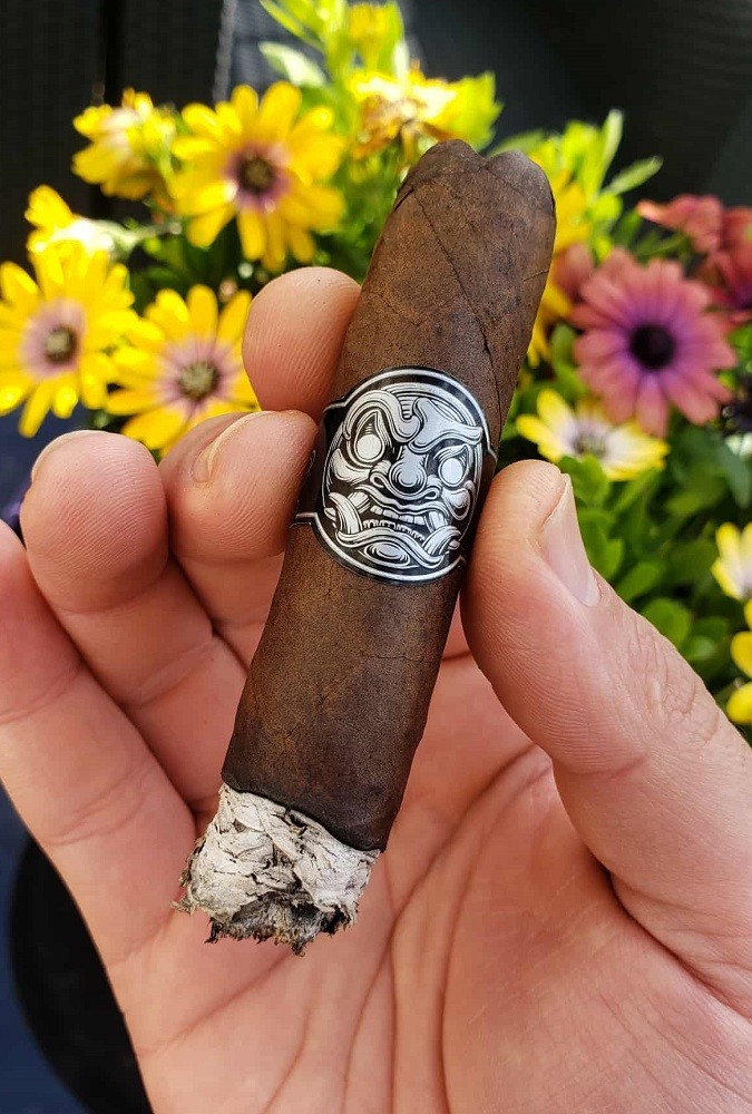 A Room 101 cigar is held. It has a medium brown wrapper and a black and white band with a samurais face on it