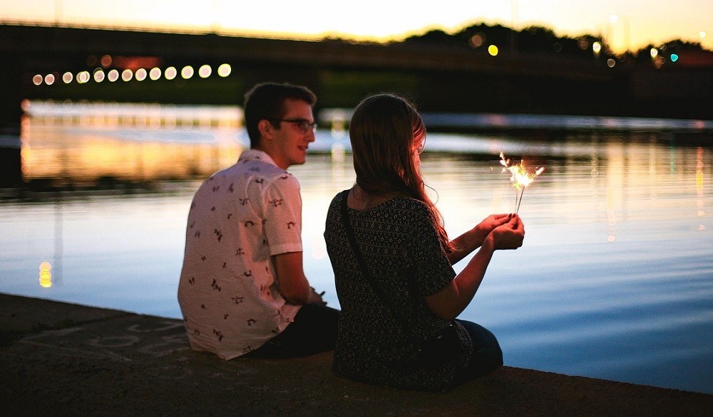 A young couple sit on the riverbank at sunset. The woman is holding a sparkler at arms length while the man stares in amazement
