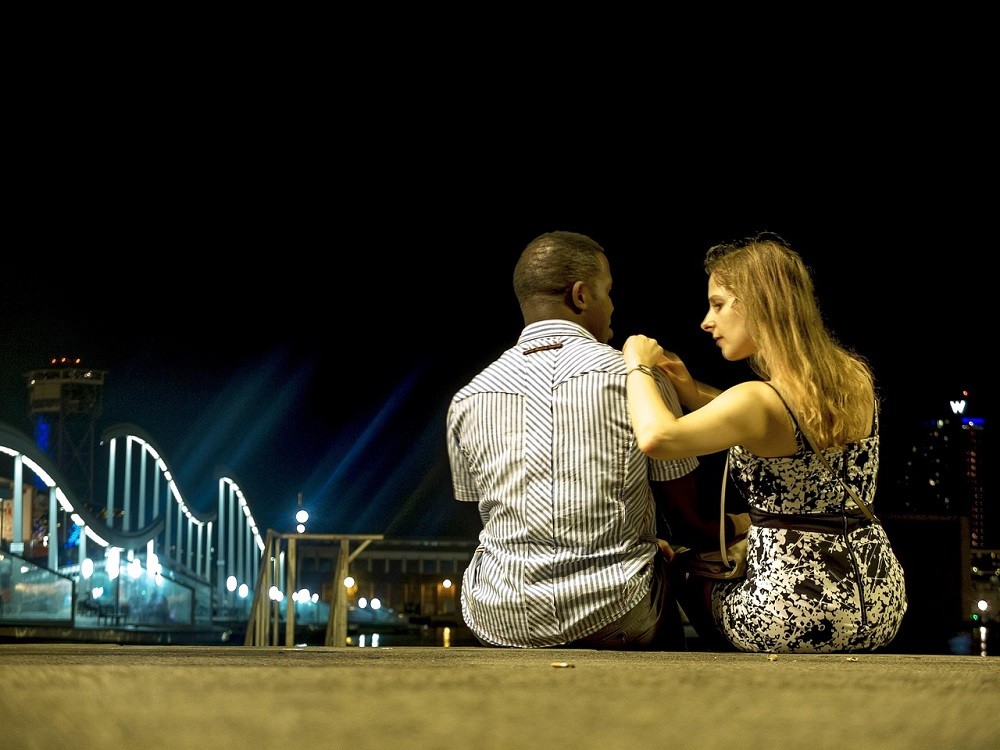 A young couple sits on a ledge at night staring at the city lights
