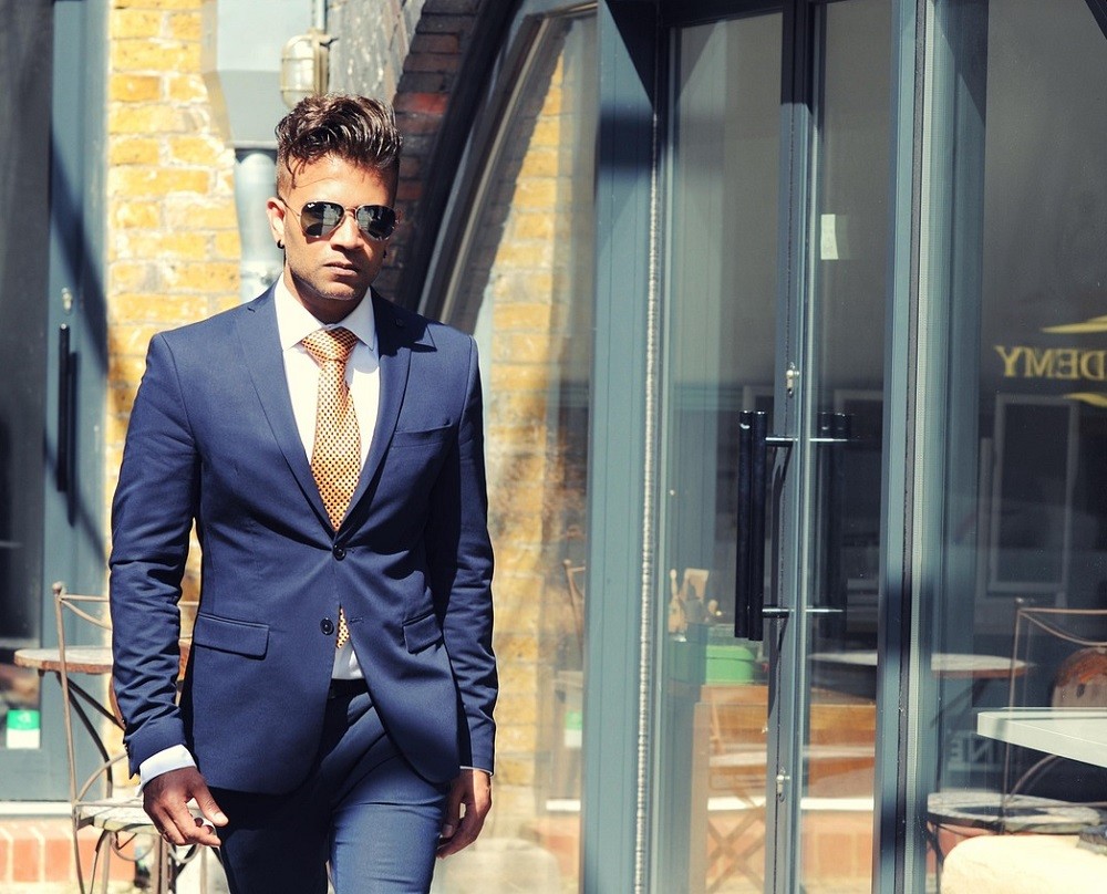 A young businessman is wearing a navy blue suit with a white shirt, gold tie, and aviator sunglasses