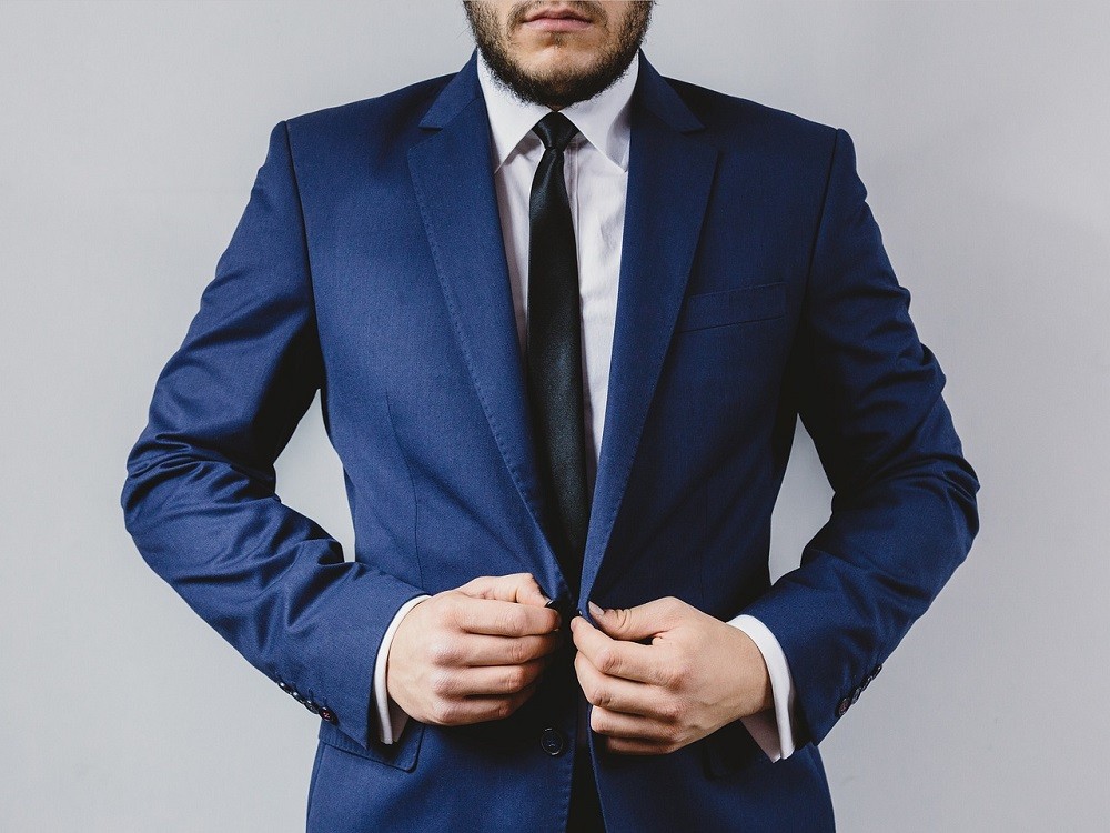 A man wearing a navy blue suit and black tie is fastening the button on his tailored suit jacket