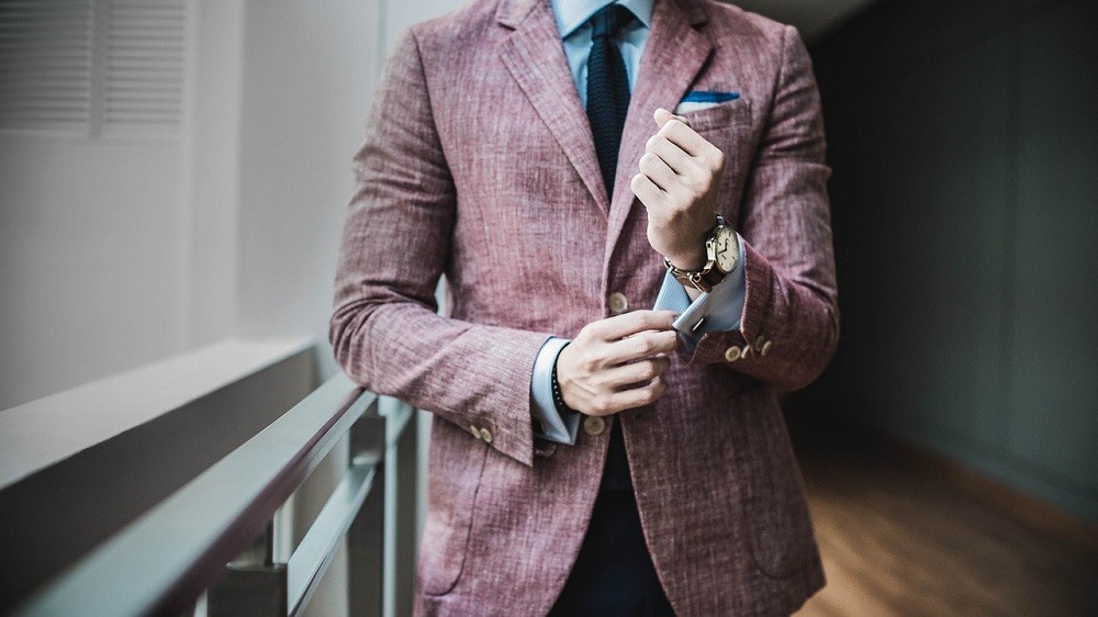 A man in a pale pink sports jacket is adjusting his cuff. He is accessorizing with a blue pocket square, black tie, and a dress watch