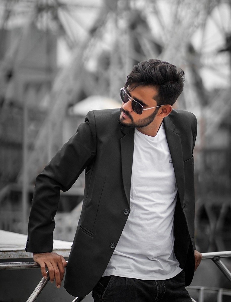 A young man with sunglasses has his charcoal grey suit unbuttoned showing off his white shirt underneath