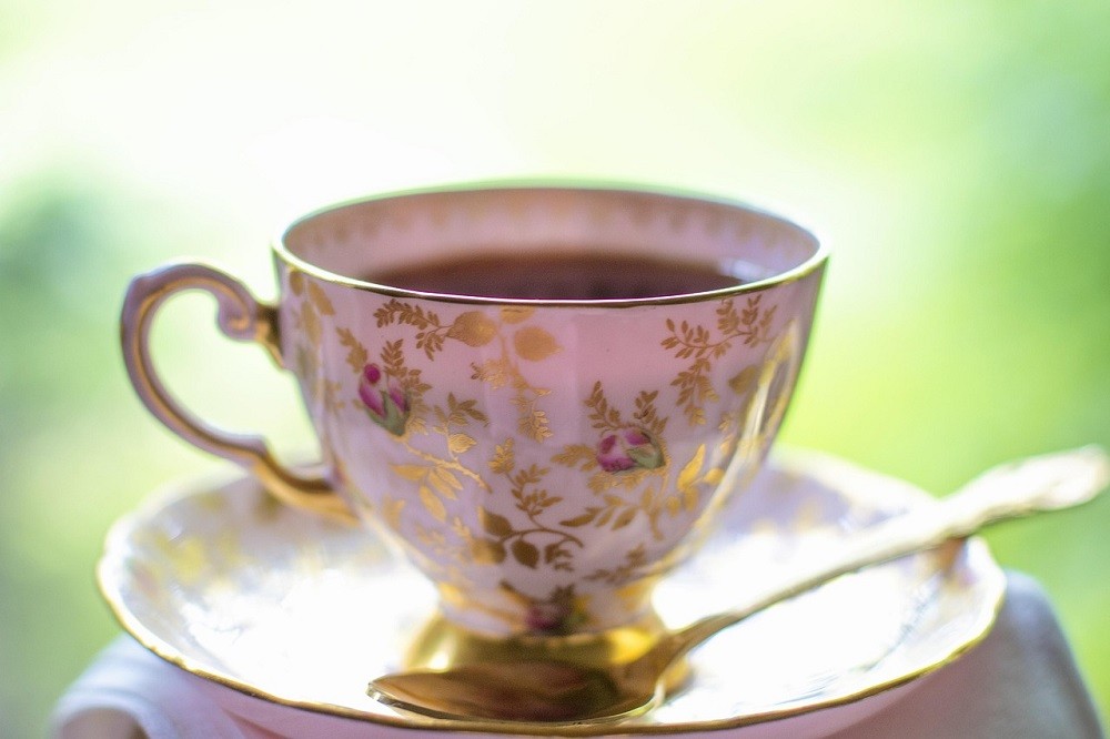 A white porcelain teacup with a gold floral pattern sits on a white saucer with a gold tea spoon
