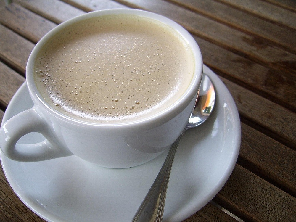A light brown latte is inside a plain white coffee mug sitting on a white saucer. There is a silver tea spoon sitting beside the mug