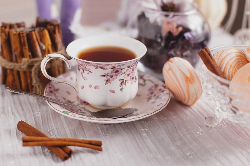 A white teacup with a purple floral pattern sits on a white saucer with the same design. A silver teaspoon rests on the saucer beside the teacup