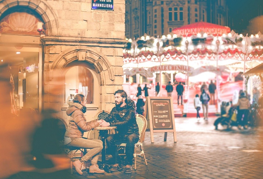 A couple sits at a small table outside a restaurant. There are people walking all around them