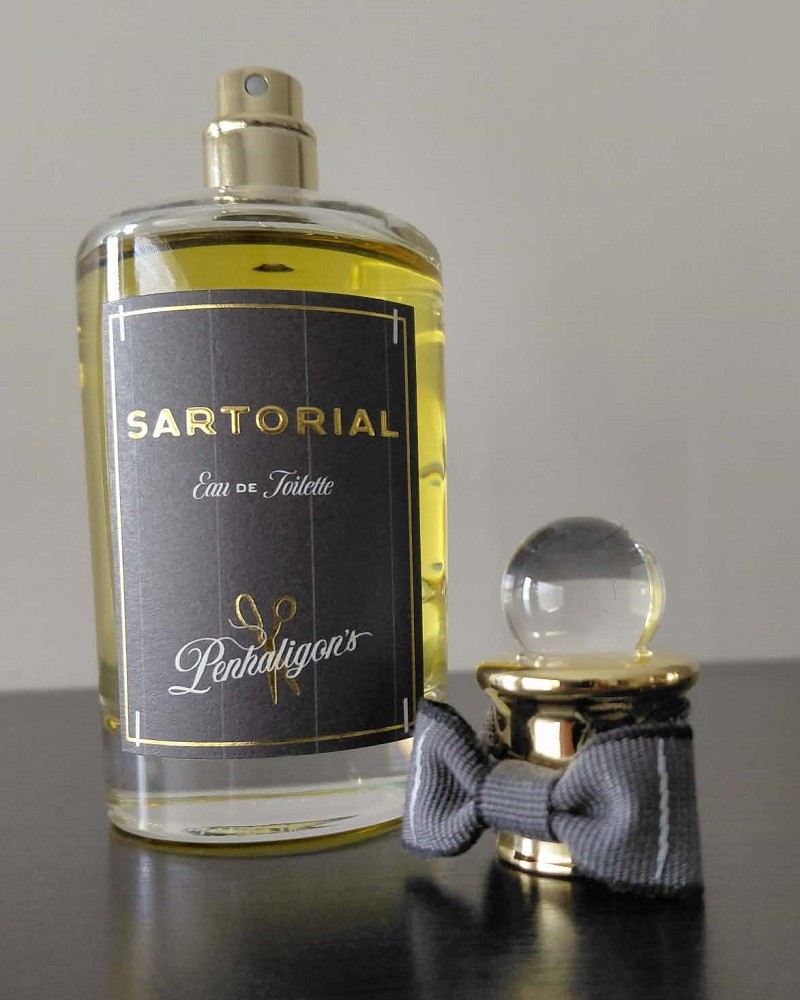A bottle of Sartorial cologne by Penhaligon's has the cap off. The cap is sitting beside the bottle. The cap has a small grey bow tie wrapped around it
