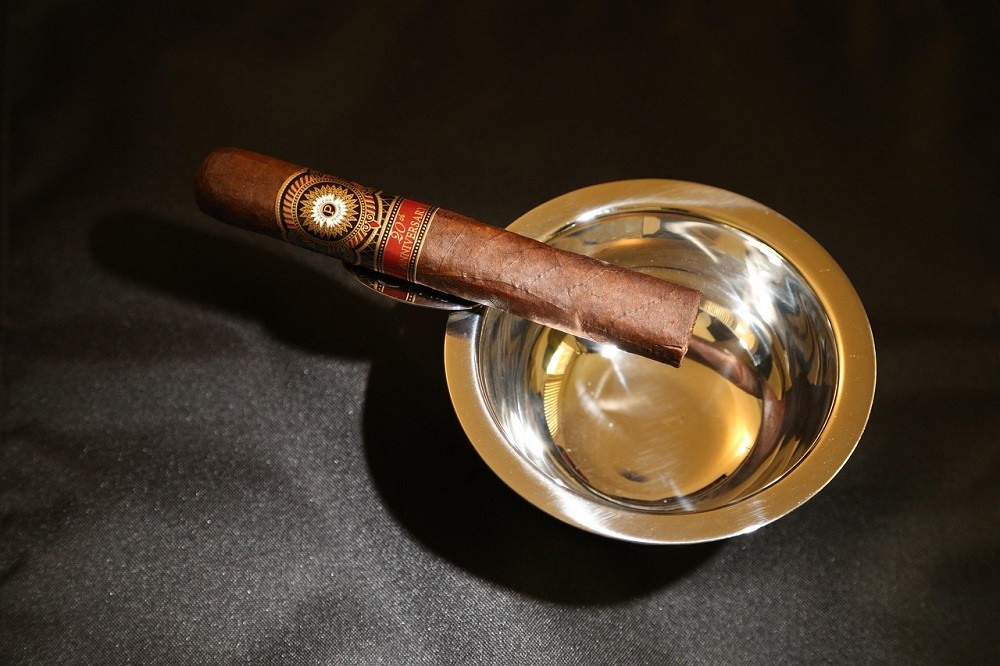 A dark brown cigar sits on the edge of a stainless steel ashtray