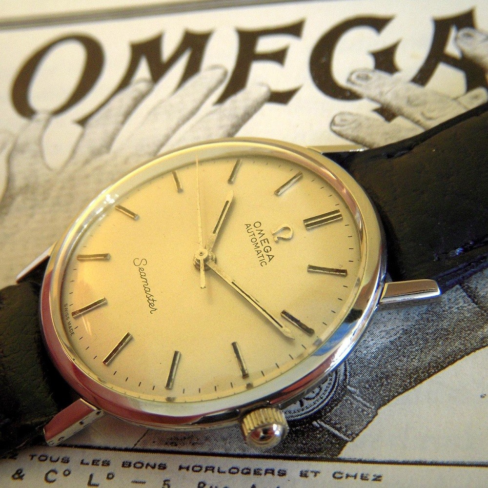 A vintage, round Omega watch with a cream-coloured dial and black strap sits on an old black and white Omega advert for the same watch