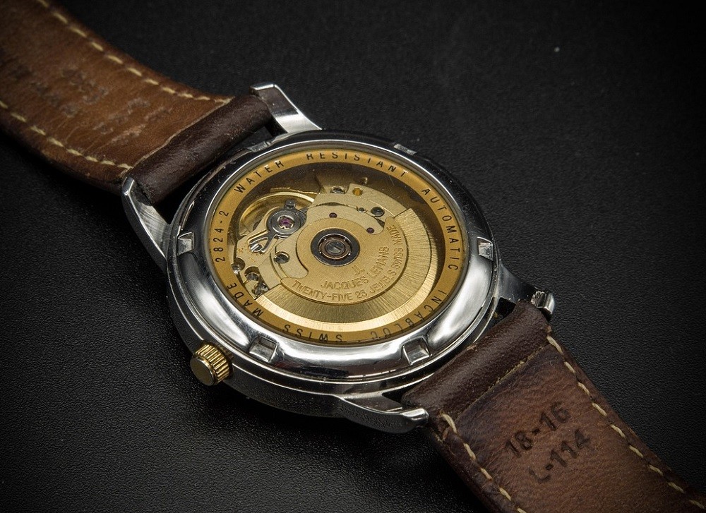 A watch sits face-down on a black table. The movement is clearly visible on the back of the watch. You can see gears, springs, levers, and all the parts necessary to make the watch tick