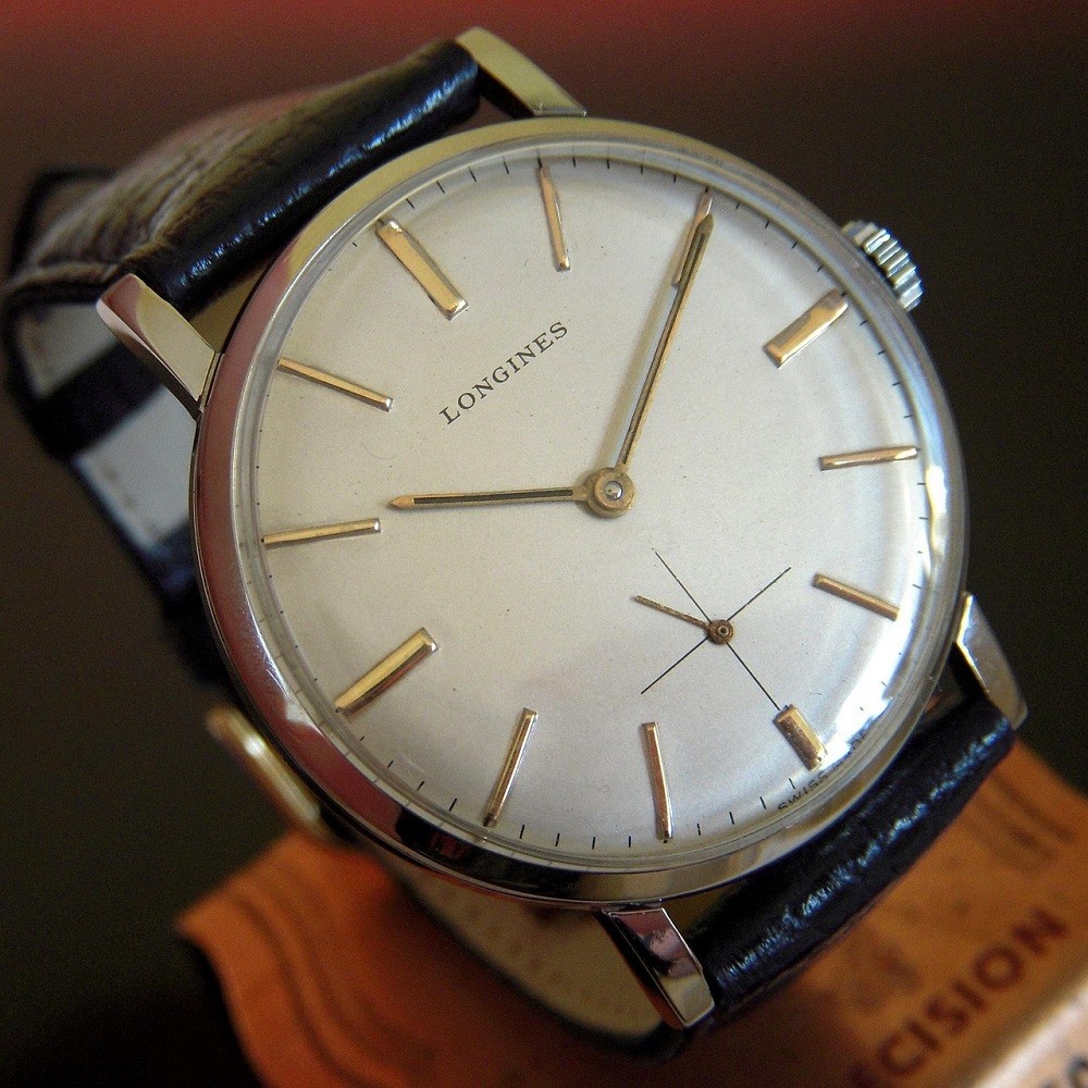 A vintage Longines watch with a white dial and gold indices is on a black strap
