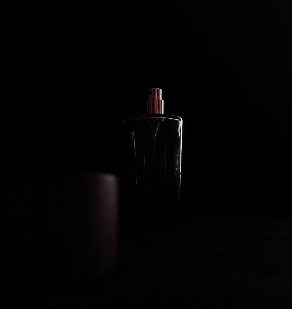 A black bottle of cologne sits on a black countertop with the only light source coming from the right side of the picture, barely outlining the bottle